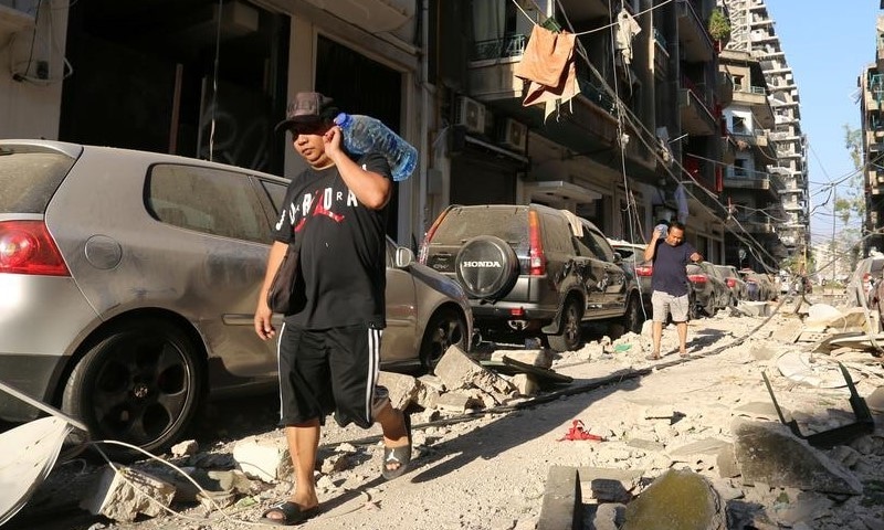 Men carrying bottles of water walk past damaged buildings and vehicles following the August 4th explosion in Beirut.